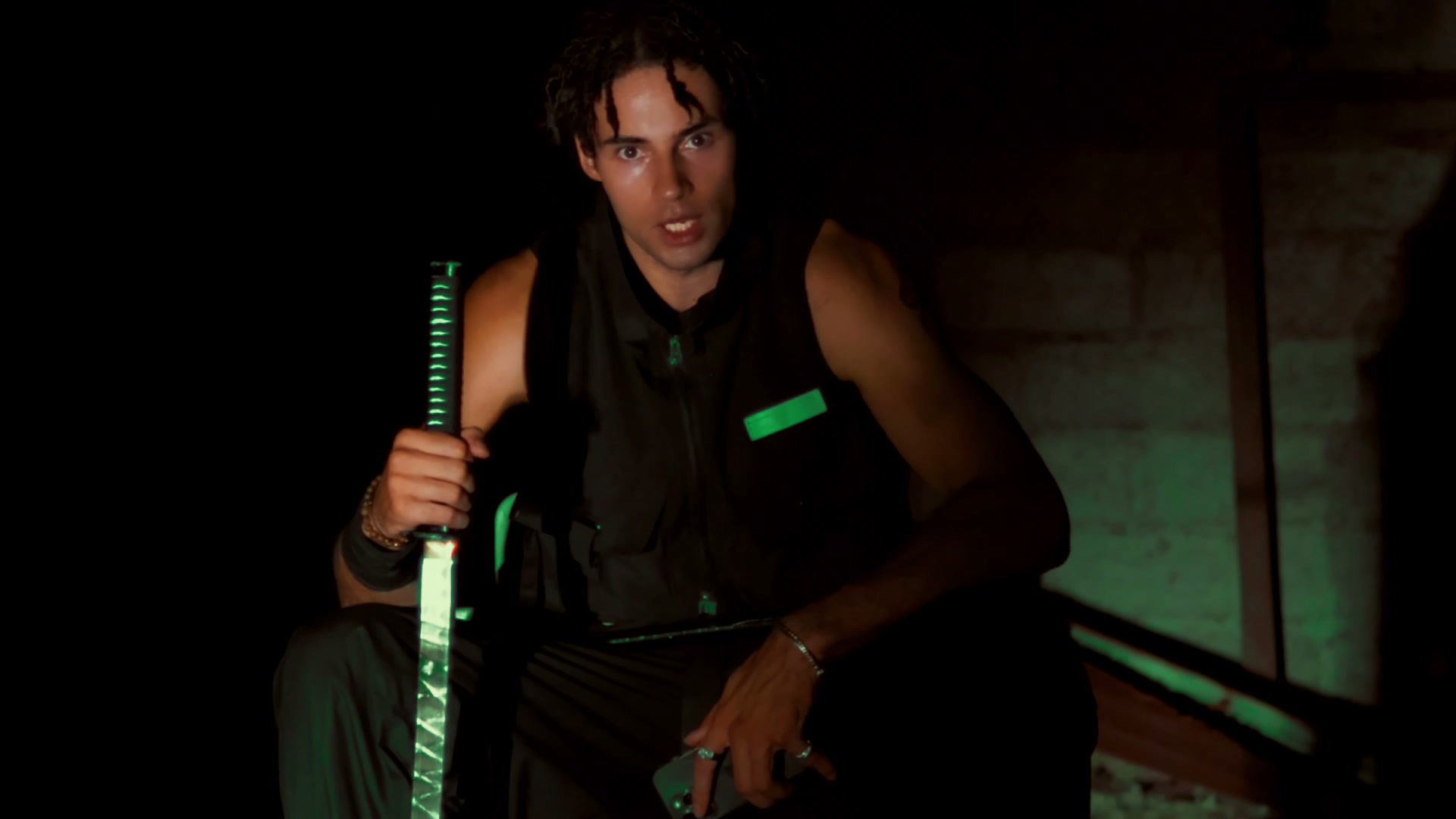 The artist crouched in a dark warehouse, gripping a katana upright with a green-lit blade, a green reflective strip across his vest catching the same light