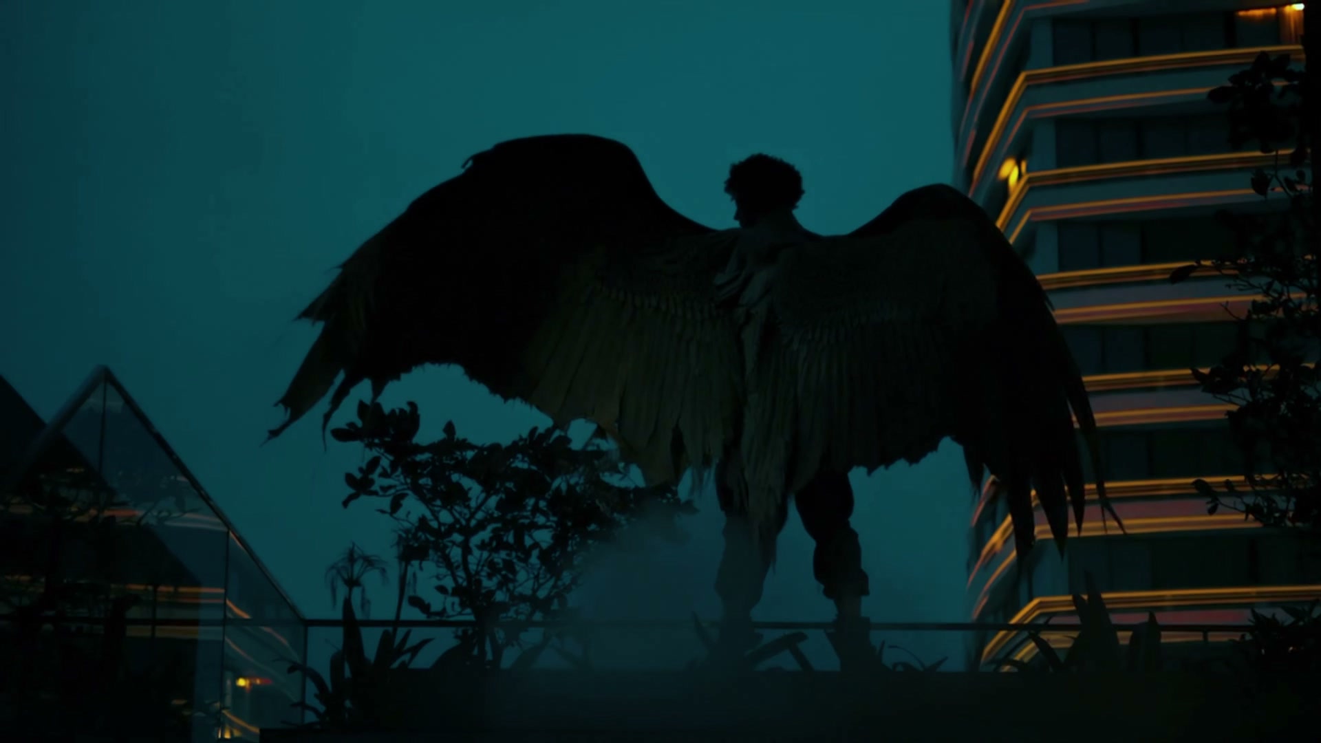A winged figure standing on a rooftop balustrade at dusk, feathered wings spread wide, silhouetted against a teal sky with a lit high-rise tower behind